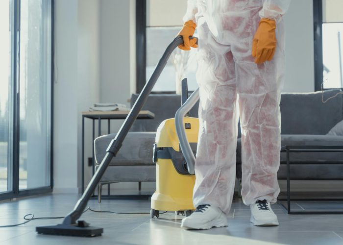A cleaner in protective coveralls and gloves vacuuming a modern living room.