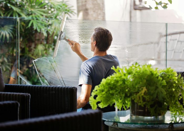 A man cleaning a glass barrier outdoors with a squeegee, surrounded by greenery.