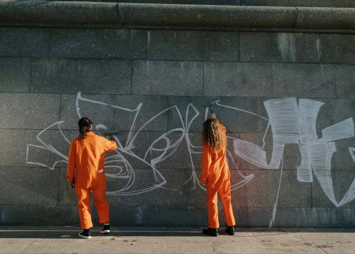 Two community service workers in orange uniforms remove graffiti from a concrete wall outdoors.